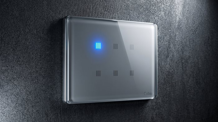 A silver glass Cubixx touch panel on a dark textured wall, one key lit blue to show the circuit is on.